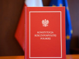Przedmiot aukcji konstytucja RP na biurku, w tle flagi Polski i Uni Europejskiej